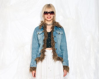 Peut inclure: Une veste en jean bleu clair avec un col et des poignets en fausse fourrure marron. La veste est portée sur un haut court noir et une jupe maxi blanche à volants. La personne porte des lunettes de soleil noires et des bottes à plateforme noires.