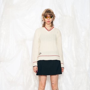 May include: A woman wearing a white sweater with a red stripe and a black pleated skirt. She is also wearing black leg warmers and sunglasses.