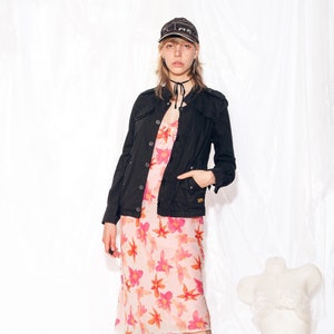 May include: A woman wearing a black jacket over a pink floral dress and black lace-up boots. She is wearing a black baseball cap with white lettering.