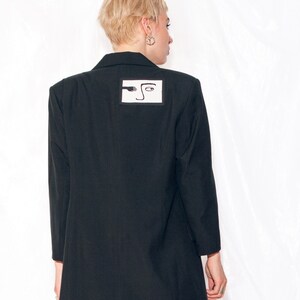 May include: A black blazer with a white patch on the back featuring a black and white abstract design of a face.