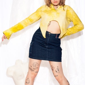 Peut inclure: Une personne portant une chemise jaune tie-dye avec une jupe en jean. La chemise est courte et a un ourlet pointu. La personne porte des chaussettes blanches et a plusieurs tatouages sur les jambes.