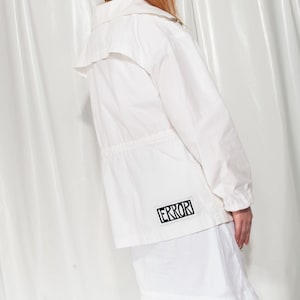 May include: White jacket with a collar and drawstring waist. The jacket has a small black and white patch that says "ERROR" on the lower back. The model is wearing a white skirt.