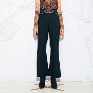May include: A woman wearing a sheer, long-sleeved top with a brown, gold and red patterned design. The top has a scoop neck and a fitted silhouette. She is also wearing black flared trousers and black heeled shoes.