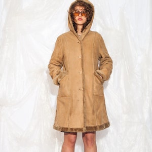 May include: A long brown suede coat with a fur-lined hood. The coat has a button closure and pockets. The model is wearing black knee-high boots.