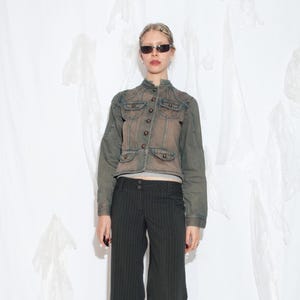 Puede incluir: Chaqueta vaquera vintage en tonos verdes y marrones, con cierre de botones, múltiples bolsillos y cuello alto. La chaqueta se combina con pantalones de raya diplomática negros. La persona lleva gafas de sol.