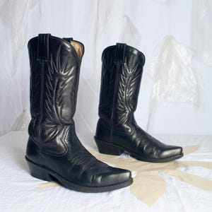 May include: A pair of black leather cowboy boots featuring a pointed toe, a stacked heel, and decorative stitching patterns on the shaft. The boots have pull tabs at the top and a smooth finish, showcasing a classic silhouette with detailed embroidery.