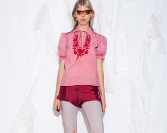 May include: A pink blouse with red floral embroidery around the neckline and sleeves. The blouse has short, puffed sleeves and a collared neckline. The model is wearing burgundy shorts and light gray leggings with knee cutouts. The model is also wearing sunglasses.