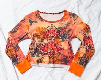Op de afbeelding: Een crop top met lange mouwen in tinten oranje, rood en zwart, met een bloemen- en scriptontwerp. De top heeft een ronde halslijn en oranje manchetten. De stof lijkt een zacht gebreid materiaal te zijn.