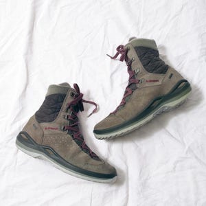 May include: A pair of brown and gray hiking boots with burgundy laces. The boots have a quilted upper and the brand name "LOWA" is visible. The boots are designed for outdoor activities and are set against a white background.