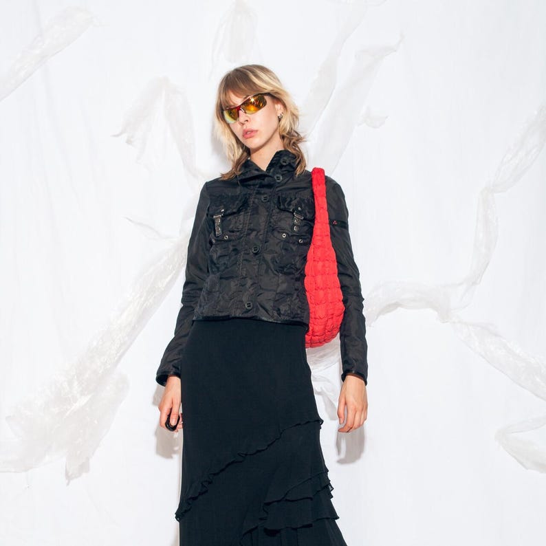 K&ouml;nnte beinhalten: Schwarze Jacke mit mehreren Taschen und Knopfleiste. Die Jacke wird mit einem langen schwarzen Rock und einer leuchtend roten Umh&auml;ngetasche kombiniert. Die Person tr&auml;gt orange get&ouml;nte Sonnenbrillen.