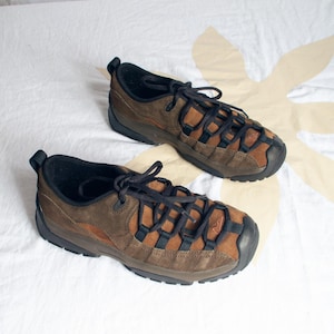 Puede incluir: Un par de zapatillas de ante marrón con cordones negros, superposiciones de malla negra, suelas de goma negra y un pequeño logotipo bordado en la puntera. El calzado de corte bajo incluye forro interior negro y tiradores en el talón.
