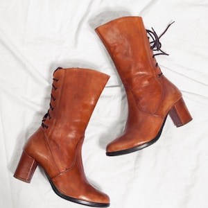 以下が含まれることがあります： 茶色の革ブーツのペア。これらのミッドカーフブーツは、背面にレースアップのデザインとブロックヒールが特徴です。ブーツは滑らかで光沢のある革で作られており、クラシックでヴィンテージなスタイルです。ヒールの高さは約7.6cmです。