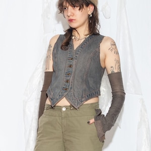 May include: A charcoal gray denim vest with a V-neckline and a front button closure, paired with long dark brown arm warmers, olive green cargo pants, and a fuzzy tan beret. The vest features a pointed hemline and visible seam detailing.