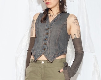 May include: A charcoal gray denim vest with a V-neckline and a front button closure, paired with long dark brown arm warmers, olive green cargo pants, and a fuzzy tan beret. The vest features a pointed hemline and visible seam detailing.