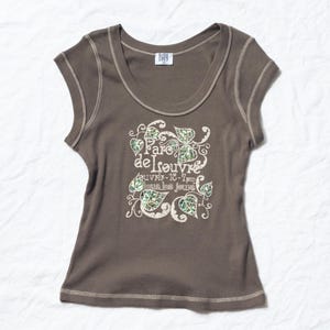Puede incluir: Una camiseta marrón, acanalada, con cuello redondo y un diseño floral blanco y verde y el texto "Parc de l'Isouvre". La camiseta tiene un ribete blanco alrededor del cuello y las sisas. La etiqueta de la marca es visible en la parte superior.