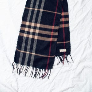 May include: A navy blue scarf with a classic plaid pattern in beige, red, and gray. The scarf has a fringed edge and a small label. The scarf is made of a soft material.