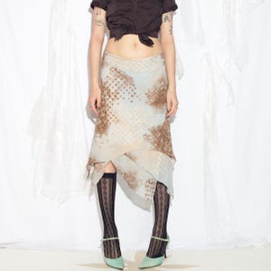 Op de afbeelding: Een rok met patroon en een asymmetrische zoom. De rok heeft een mix van bruine en witte stippen en een lichtblauwe achtergrond. De outfit bevat zwarte kousen met patroon en mintgroene hakken.