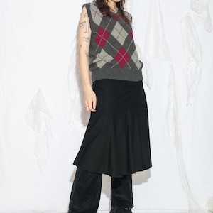 May include: A person wearing a gray argyle sweater vest with red diamond accents, a black skirt, and black platform shoes. The outfit is set against a white backdrop.