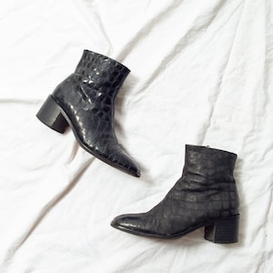 May include: Two black ankle boots with a textured, crocodile-effect pattern. The boots feature a pointed toe and a block heel, approximately 5 cm high. The footwear is displayed on a white, creased fabric background.