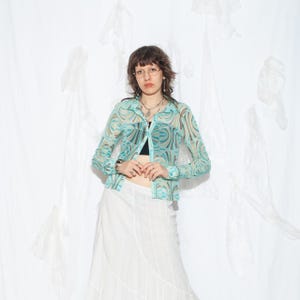 May include: A sheer, turquoise button-down shirt with a brown and turquoise swirl pattern. The shirt is paired with a black top and a white skirt. The outfit is styled with silver jewellery and glasses.