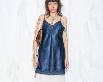 May include: A navy blue satin slip dress with lace detailing. The dress features spaghetti straps and a V-neck. A brown shoulder bag with a gold chain is visible. The person is wearing a silver bracelet and multiple necklaces.