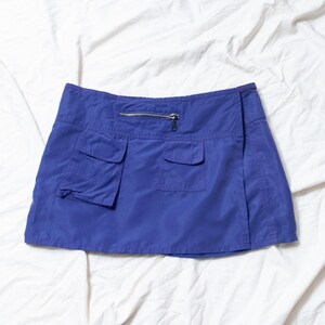 May include: A bright blue mini skirt featuring a zip and two front pockets. The skirt is made from a smooth, lightweight material. It has a classic, casual design.