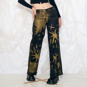 Puede incluir: Pantalones negros y dorados estampados con cintura alta. Los pantalones presentan un diseño único con motivos de sol y estrellas. La tela tiene un efecto tie-dye en oro y negro. Los pantalones tienen una pernera acampanada y se llevan con zapatos negros.