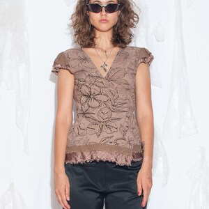 Puede incluir: Una blusa marrón de manga corta con un estampado floral de líneas en marrón oscuro, escote en V y un dobladillo con volantes y sutiles lentejuelas, combinada con pantalones de satén negro. La blusa tiene mangas casquillo y diseño cruzado.