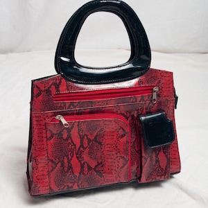 Peut inclure: Un sac à main rouge avec un motif peau de serpent noir, doté d'une anse arquée noire brillante, d'une poche frontale zippée et d'une petite poche à rabat noire sur le côté droit. Le sac présente des fermetures éclair argentées et une forme rectangulaire.