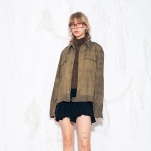 Puede incluir: Chaqueta verde oliva con dos bolsillos en el pecho y cierre de botones. La chaqueta se lleva sobre un jersey de cuello alto marrón y se combina con una falda negra. La persona lleva gafas.