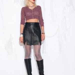 Könnte beinhalten: Eine Frau trägt ein lilafarbenes Strick-Crop-Top, einen schwarzen Leder-Minirock und schwarze kniehohe Stiefel.