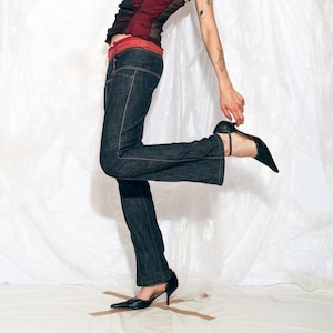 Puede incluir: Vaqueros de mezclilla de lavado oscuro con costuras naranjas y un ligero acampanado. Los vaqueros se combinan con un top negro y tacones negros de punta. El conjunto se encuentra sobre un fondo blanco.