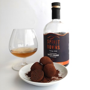 May include: A bottle of Sussex Brandy with a dark label, the text "Spirit of the Downs", and a snifter glass with amber liquid. A small white plate holds chocolate truffles dusted with cocoa powder.
