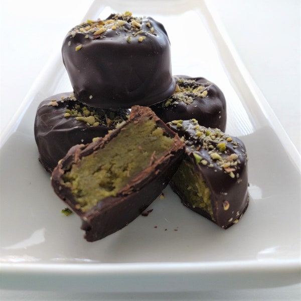 May include: Close-up of several dark chocolate-covered confections, some whole and some cut open to reveal a pistachio filling. The treats are topped with chopped pistachios and arranged on a white rectangular plate.