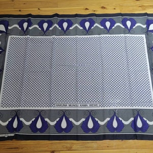 Può includere: Un tessuto bianco con un bordo nero e un motivo blu e bianco. Il tessuto presenta un motivo ripetitivo di forme a goccia con un centro bianco e un contorno blu. Il tessuto presenta anche un motivo a scacchi bianco e nero al centro. Il tessuto ha il testo "Hakuna Mungu Kama Wewe" stampato in nero.