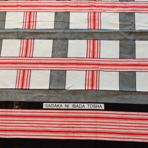 May include: White fabric with a red and black checkered pattern. The fabric has the text "SADAKA NI IBADA TOSHA" printed on a black label.