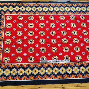 May include: Red fabric with a black and yellow border and a repeating pattern of concentric circles. The fabric has the text "Mungu ni Muweza wa Yote" printed in black.