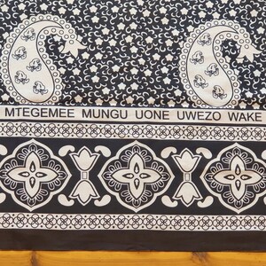 May include: Black and white fabric with a paisley pattern and the text "MTEGEMEE MUNGU UONE UWEZO WAKE". The fabric has a border with a repeating pattern of white flowers and geometric shapes.