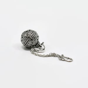 May include: A silver charm with a delicate, intricate design. The charm is shaped like a sphere and has a small chain attached to it.