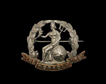 Op de afbeelding: Zilverkleurige broche met een zittende figuur met een speer en een schild met de Union Jack. De figuur is omgeven door een lauwerkrans, met een banner eronder met de tekst "THE NORFOLK REGT."