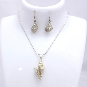 Könnte beinhalten: Ein silbernes Halsketten- und Ohrring-Set mit einem detaillierten Muschel-Anhänger und passenden Ohrringen. Die Halskette hat eine zarte Kette und der Anhänger ist eine realistische Darstellung einer Muschel.