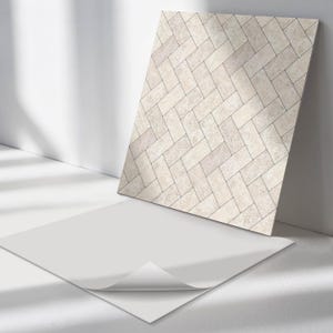 May include: A rectangular tile with a herringbone pattern in shades of beige and grey, leaning against a white wall. A white sheet of paper with a curled corner is in the foreground.