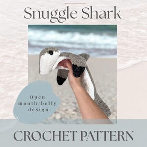 Könnte beinhalten: Ein Häkelmuster für einen Snuggle Shark mit offenem Mund/Bauch-Design. Der Hai ist grau, weiß und schwarz, mit dem Text "COLDWATER SHORES" und "CROCHET PATTERN". Der Hintergrund zeigt eine Strandszene.