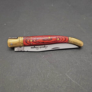 May include: A small, red and black pocket knife with a brass handle and a curved blade. The handle is decorated with gold dots and the blade is engraved with the words "Laguiole Tradition".