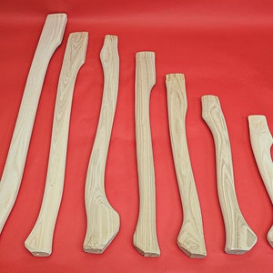 Könnte beinhalten: Acht unfertige Holz-Axt-Stiele in verschiedenen Längen, in einer Reihe auf rotem Hintergrund angeordnet.