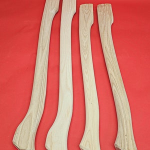 Può includere: Quattro manici di ascia in legno grezzo, tutti con una forma e una venatura simili. I manici sono disposti in fila su una superficie rossa.