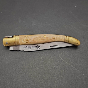 May include: A small, folding pocket knife with a wooden handle and brass accents. The blade is partially open and the handle has a brass inlay with a pattern of dots. The knife is engraved with the words "Sapinero Secateur".