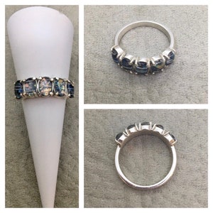 May include: A silver ring with a row of oval-shaped iridescent gemstones. The ring is on a white display stand.