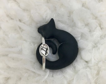 Puede incluir: Un anillo de plata con una piedra ovalada transparente engastada en una banda de plata. El anillo se muestra en una figurita de gato negro.