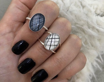 May include: Two silver rings with oval settings featuring embedded black hair strands. One ring has a dark grey background with a twisted band, while the other has a white background with a smooth band. Both display a crisscross pattern of hair.